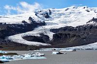 Ableger  Da macht man schon ein Panorama, mangels eines sehr kurzbrennweitigen Objektivs bestehend aus einigen Hochkantaufnahmen und schafft es trotzdem nicht, den gesamten Gletscher vernünftig auf einer Bildschirmseite abzubilden. Deshalb gibt es hier die linksseitige Fortsetzung des vorigen Bildes, den durch einen Hügelzug vom Hauptgletscher abgetrennten Ableger des Fjallsárlóns. Wie ich im Internet erfahre, war bis vor 16 Jahren die schmale Zunge am Fuße des Hügels noch mit dem Hauptfeld vereint. Heute endet er bereits etwa einen Kilometer vor dem Hauptfeld und hat einen eigenen kleinen Schmelzwassersee, von dem der Überlauf als schmales Rinnsaal im großen See mündet.  Nachdem wir noch etliche Fotos von den, teils als durchsichtige Brocken herumtreibenden Eisschollen geschossen haben, führt uns der Rundweg wieder zum Parkplatz zurück.