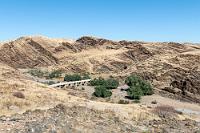 Kuiseb Pass  Die Straße senkt sich nun schnell hinab zum Kuiseb Pass, einer Brücke über den knochentrockenen Flusslauf des westlich Windhuks entspringenden Kuiseb Rivers, dessen Oberlauf die Niederschläge des Khomashochlandes aufnimmt, dann die Grenze zwischen der Wüstennamib und der Dünennamib markiert, um schließlich nach ca. 560 km südlich von Walvisbay in den Atlantik zu münden. Dies passiert allerdings durchschnittlich nur alle 10 Jahre, so dass der Fluß in der Regel obiges Erscheinungsbild zeigt.